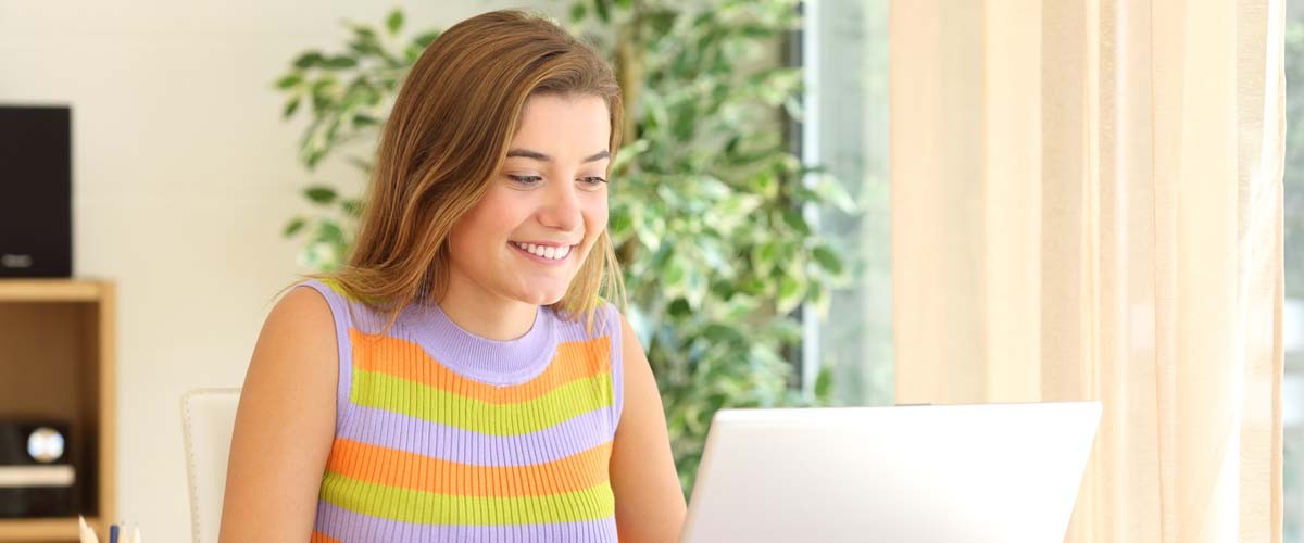 Portrait of a casual student studying on line using a laptop sitting in a chair in a table at home with a window in the background